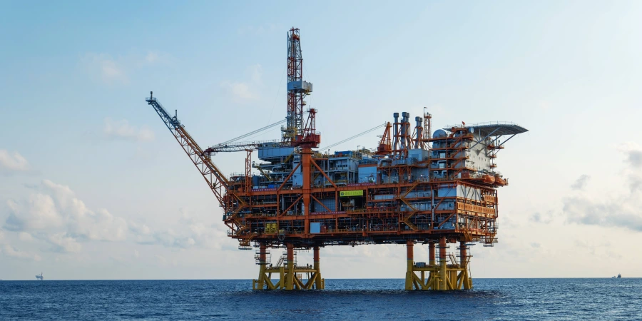 Large orange offshore oil platform standing on yellow steel legs in calm blue ocean waters under a clear sky, featuring multiple deck levels, tall drilling derrick, cranes, helipad, and extensive piping and equipment.