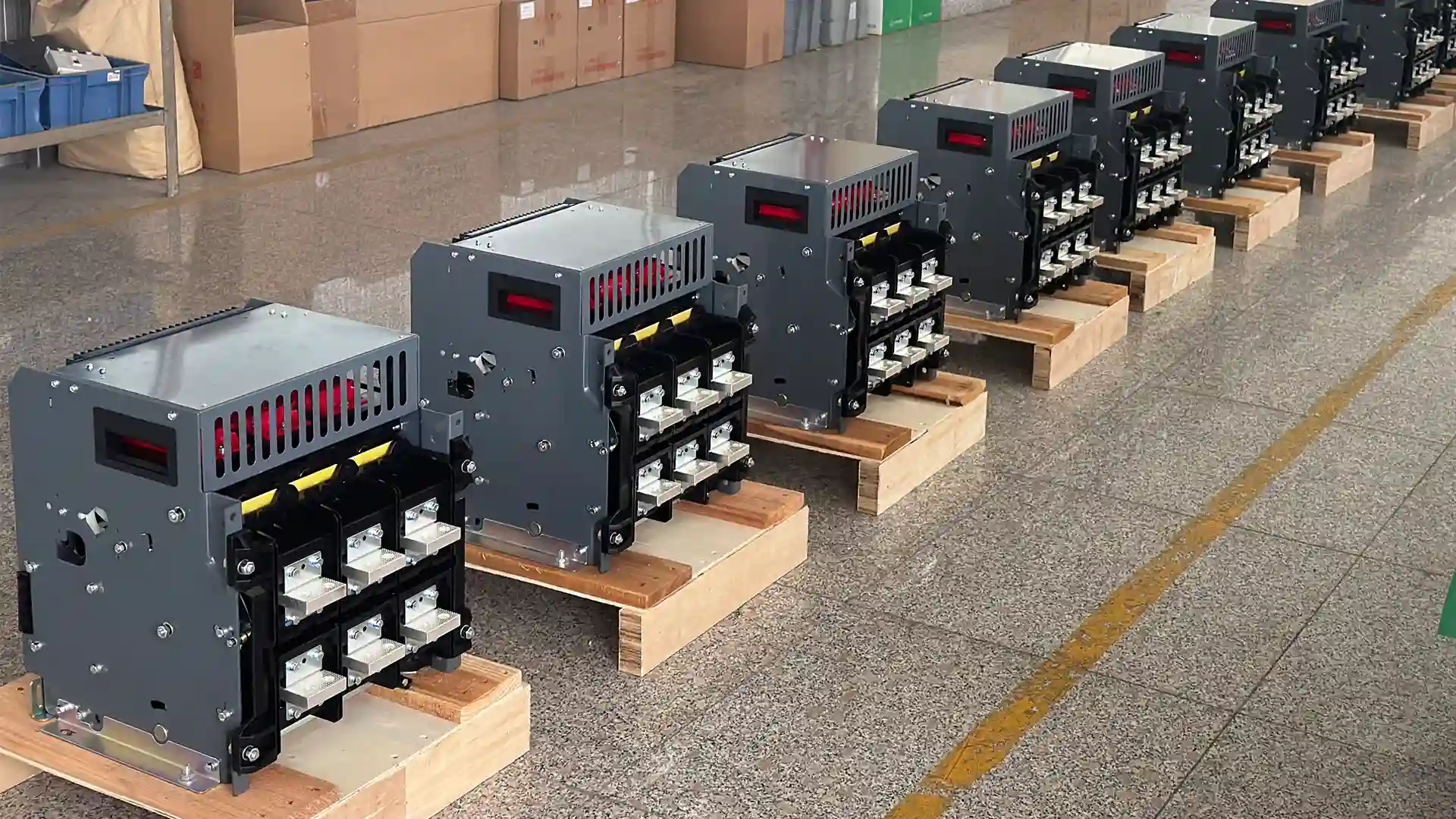 Multiple gray air circuit breakers (ACBs) arranged in a neat row on wooden pallets inside a workshop or factory assembly area. Each unit features ventilation slots, exposed busbar terminals with white connectors, red internal components visible through openings, and a clean industrial design, with cardboard boxes and equipment in the background.