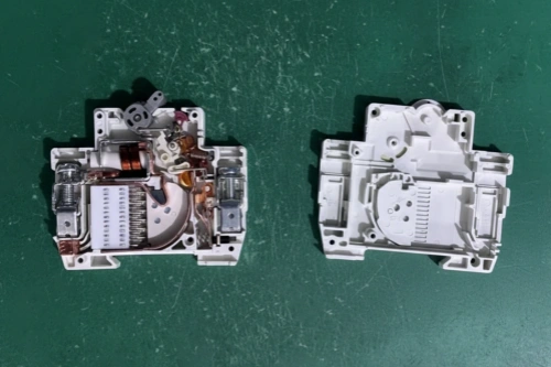 Two white plastic housings of a miniature circuit breaker (MCB) placed side by side on a green surface. On the left is the semi-assembled SKD unit showing internal components including the electromagnetic coil, bimetallic strip, arc chute, contacts, and trip mechanism. On the right is the empty housing displaying the internal structure and mounting points without components.