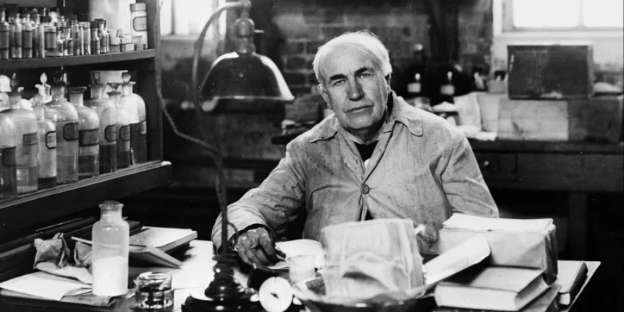 A black-and-white photo of inventor Thomas Edison sitting at a desk in his lab. He has white hair and is wearing a light jacket, looking at the camera. The desk is cluttered with books, papers, glass bottles, and lab equipment. Shelves behind him hold many more bottles, and a large lamp hangs overhead.