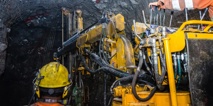 Two workers in orange high-visibility gear and yellow hard hats operate a large yellow twin-boom electro-hydraulic jumbo drill rig inside a dark underground mine tunnel with rock walls reinforced by mesh netting.