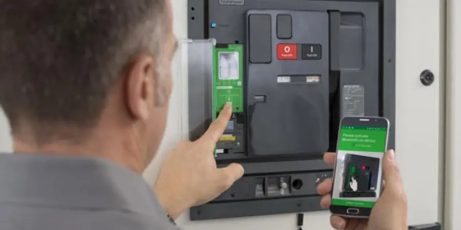 A technician viewed from behind, pointing to a communication module inserted into a smart air circuit breaker or molded case circuit breaker mounted in an electrical panel. The module features a green circuit board, while the breaker has red 'Alarm' and black 'Test' buttons. In the other hand, the technician holds a smartphone displaying a green app interface with instructions for Bluetooth activation and remote control.