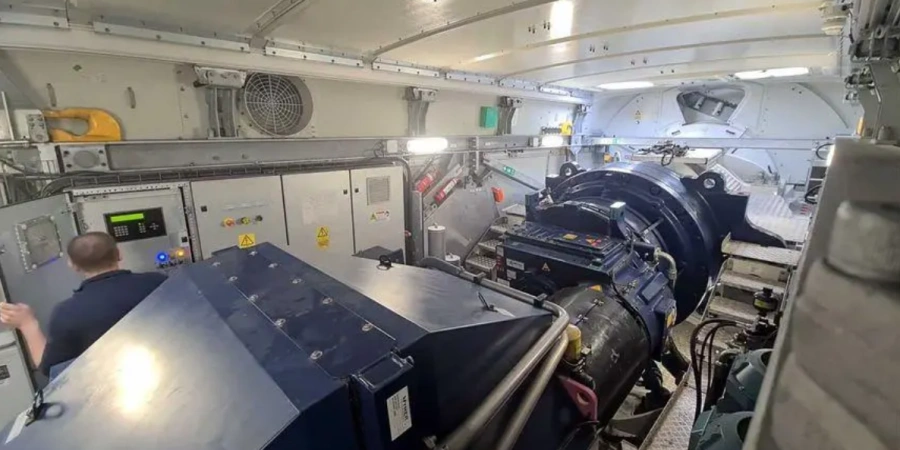 Interior view of a wind turbine nacelle showing the large blue gearbox and generator assembly, surrounded by control cabinets, cables, cooling fans, and a technician working in the confined, brightly lit space high above the ground.