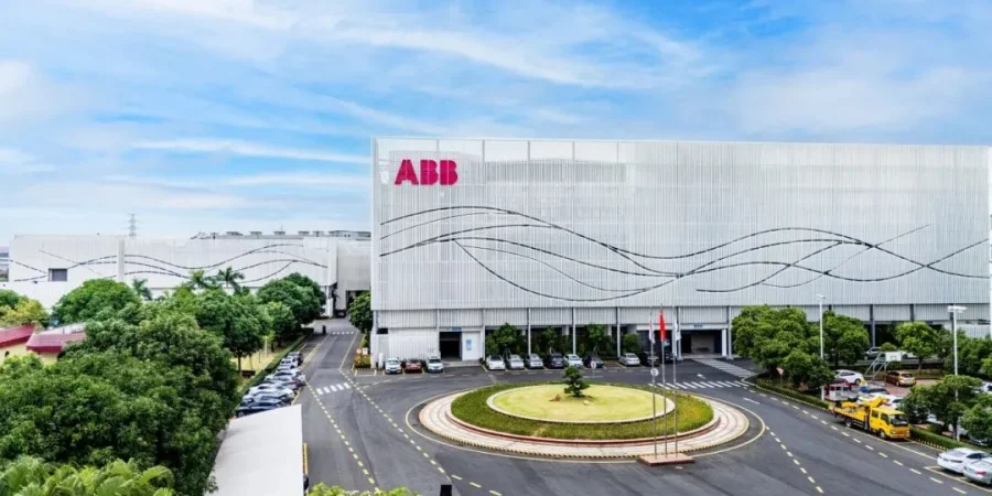 Modern white industrial building with large red ABB logo on the facade and wavy black line designs, surrounded by green trees, a roundabout with parked cars, and a clear blue sky in Xiamen