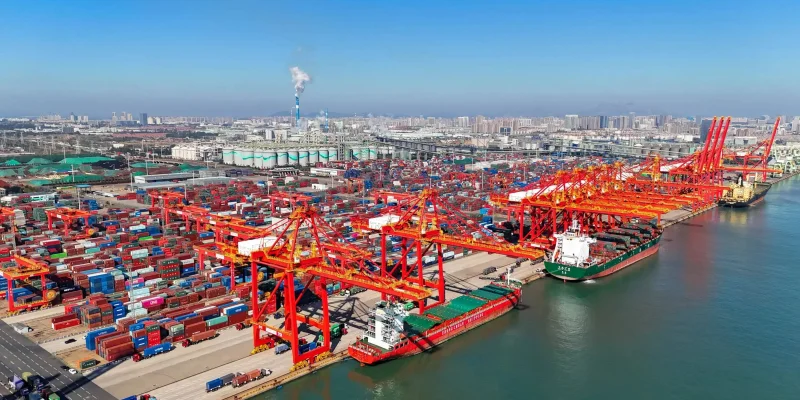 Drone aerial view of a massive container terminal on a clear day, filled with thousands of multicolored shipping containers stacked in organized rows, several large red gantry cranes actively working along the docks, a green cargo ship being loaded at the pier, industrial tanks and chimneys in the distance, and a city skyline visible under bright blue skies.