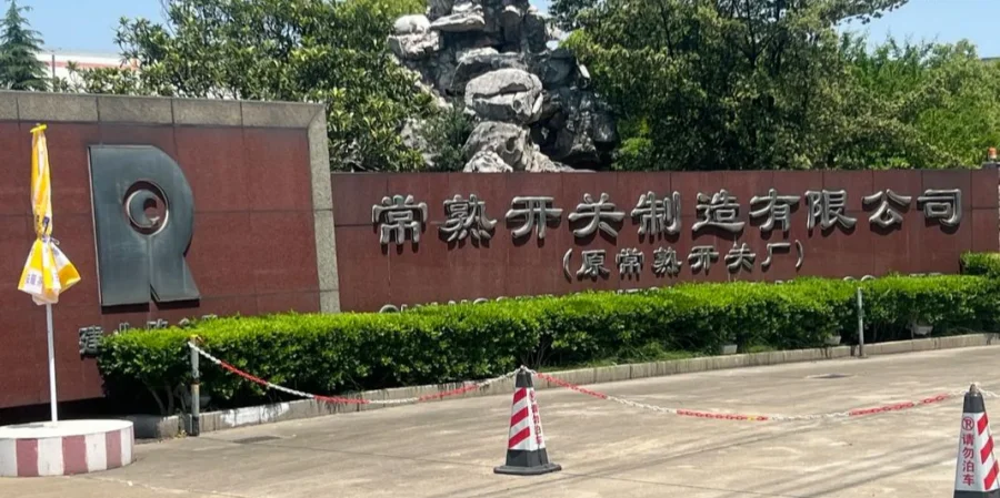 Front gate sign of Changshu Switch Manufacturing Co., Ltd. (formerly Changshu Switch Factory) in Chinese characters on a red wall, with a large R logo, green hedges, and a rock sculpture in the background
