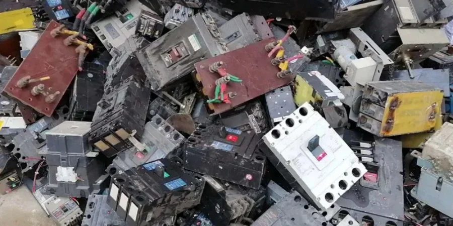 A large heap of old and used electrical circuit breakers piled together. The breakers are mostly black, gray, and white, some with colorful wires attached and visible rust or dirt, showing they are scrap or waste ready for recycling.