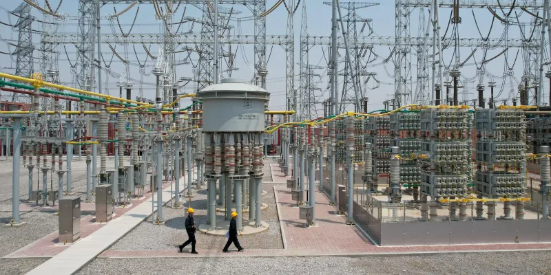 Wide view of a large outdoor electrical substation on a sunny day, featuring tall metal lattice structures supporting thick overhead power lines, numerous gray transformers and insulators, colorful yellow and green insulated busbars and cables, large cylindrical equipment, and two workers in yellow hard hats and dark uniforms walking along a gravel path between the equipment.