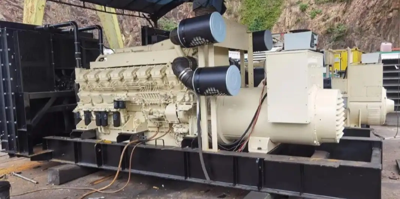 Large industrial diesel generator mounted on a black metal base frame in an outdoor workshop or construction site. The unit features a beige engine block with six visible cylinders, black air intake pipes with blue covers, an attached alternator at the end, various hoses and cables, exhaust components, control boxes, and surrounded by rocky cliff background under a shaded canopy.