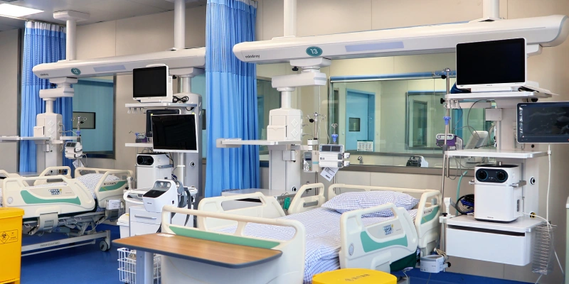 A clean, modern hospital ward showing multiple patient beds with blue privacy curtains, ceiling-mounted articulated arms holding monitors and medical devices, flat-screen displays, IV poles, ventilators, and other equipment. The beds have green and white bedding, and the floor is blue with yellow medical waste bins visible.