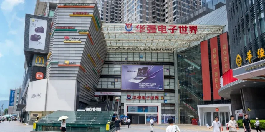 Front entrance of a large modern shopping mall named Huaqiang Electronics World in Shenzhen's Huaqiangbei district, with colorful building facade, brand ads for Xiaomi, Huawei, and Dell, and people walking outside under a covered walkway.