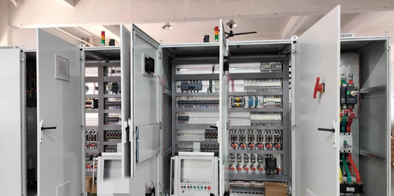 Several large grey metal electrical control cabinets with open doors, revealing neatly organized internal components. Visible inside are rows of circuit breakers, terminal blocks, relays, wiring, contactors, and control modules. Red and green indicator lights, emergency stop buttons, and a small control panel with switches are mounted on the front. The setup is installed in an industrial indoor space with concrete floor and ceiling fans.