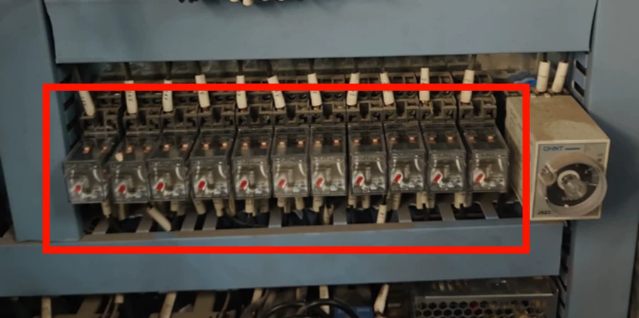 Row of multiple gray intermediate relays mounted on a DIN rail inside a blue electrical control panel, each with transparent covers showing red indicator flags, screw terminals, and wiring connections, next to a timer relay.