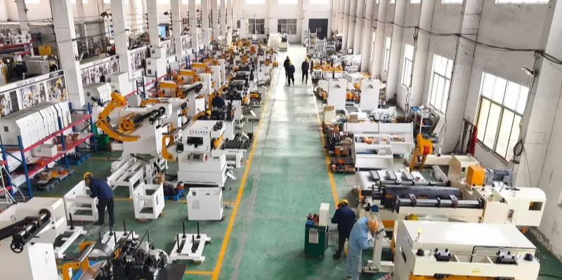 A very large factory workshop with high ceilings and rows of various metalworking machines, including lathes, milling machines, and CNC equipment. Workers in helmets and blue uniforms are operating machines along the green floor with yellow walkway lines. The space is filled with white and orange machinery, tools, and workstations under bright indoor lighting.
