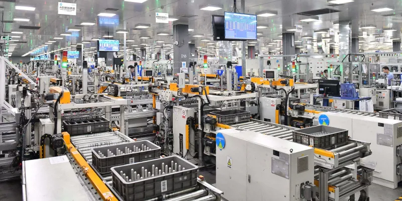 Wide interior view of a large, bright factory floor with automated production lines. Rows of complex conveyor systems, robotic arms, orange and white machinery, conveyor belts carrying gray plastic crates filled with test tubes or vials, overhead monitors displaying data, workers in blue uniforms visible in the background, and bright ceiling lights illuminating the high-tech assembly environment.