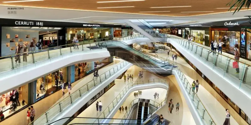 A spacious, multi-storey shopping mall interior with elegant curved escalators connecting several floors. The design features smooth white and beige architecture, glass railings, warm wooden ceiling panels, and bright overhead lighting. Shoppers walk along the walkways, while luxury and fashion brand stores.