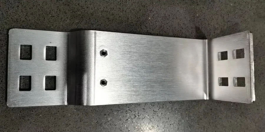 A single nickel-plated metal busbar with a bent shape, featuring four square holes on each end and two central bolts, placed on a gray speckled surface