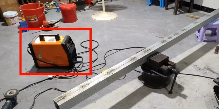 Small orange Dongcheng inverter welder with black handle placed on a concrete workshop floor next to a long metal beam clamped in a vise with welding cables connected
