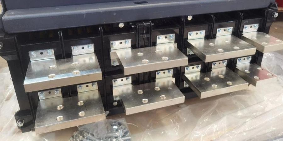 Close-up of the rear terminal section of a 4-pole air circuit breaker (ACB), showing four sets of silver-plated copper connection pads or busbar terminals arranged in a row. Each pole has multiple drilled holes for bolting cables or busbars, mounted on black insulating supports with visible mounting brackets and hardware. The breaker housing is dark gray at the top, and small bags of bolts and nuts are visible in the foreground on plastic wrapping in a workshop setting.