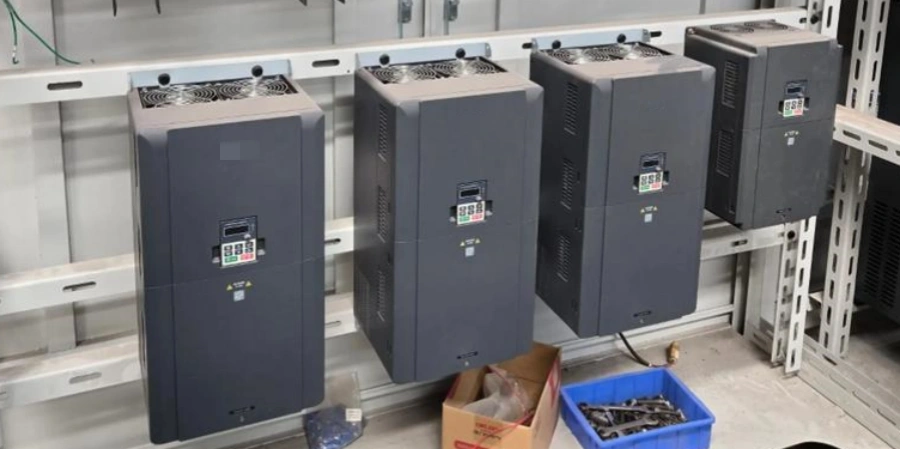 Four tall grey variable frequency drives (VFDs) mounted side by side on a metal rack in an industrial setting. Each has cooling fans on top, small display panels, and warning labels, with some units stacked in pairs. Below are cables, a blue plastic crate, and a cardboard box containing tools.