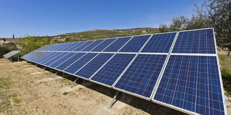 A large set of blue solar photovoltaic panels mounted on metal frames, installed on a grassy hillside. The panels are arranged in rows under a clear blue sky, with green olive trees, rolling hills, and a small village visible in the background.