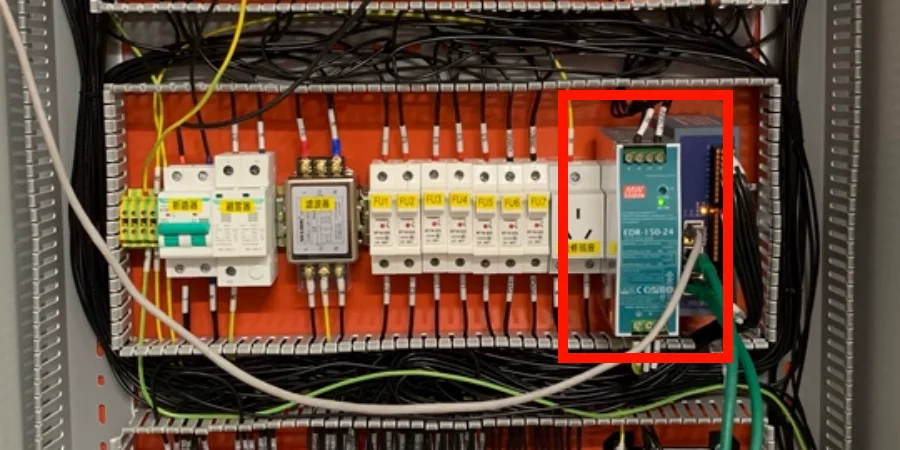 Close-up of a slim blue and green switched-mode power supply unit mounted on an orange DIN rail inside an electrical control panel, with multiple connection terminals, and surrounding fuses, circuit breakers, and bundled cables