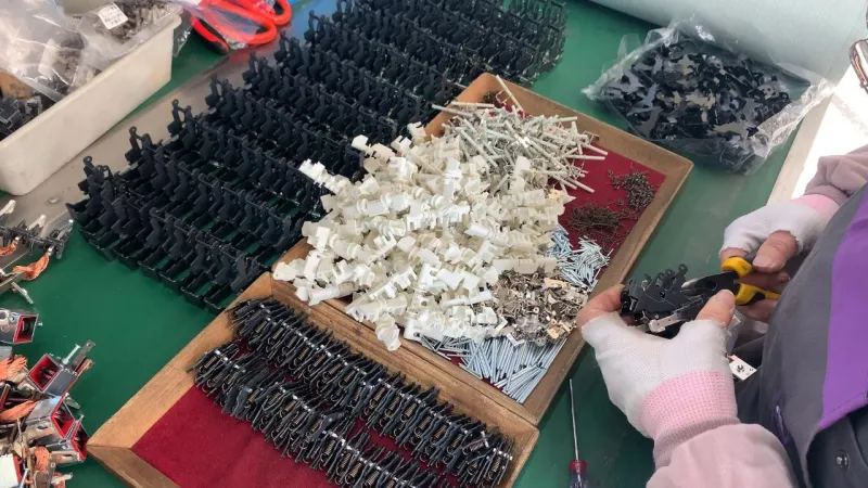 Person wearing white and pink gloves using yellow-handled pliers to assemble black plastic components of an MCCB on a green workbench surrounded by neat rows of black multi-pole circuit breaker parts piles of white insulating pieces small metal screws and other hardware in trays and plastic bags