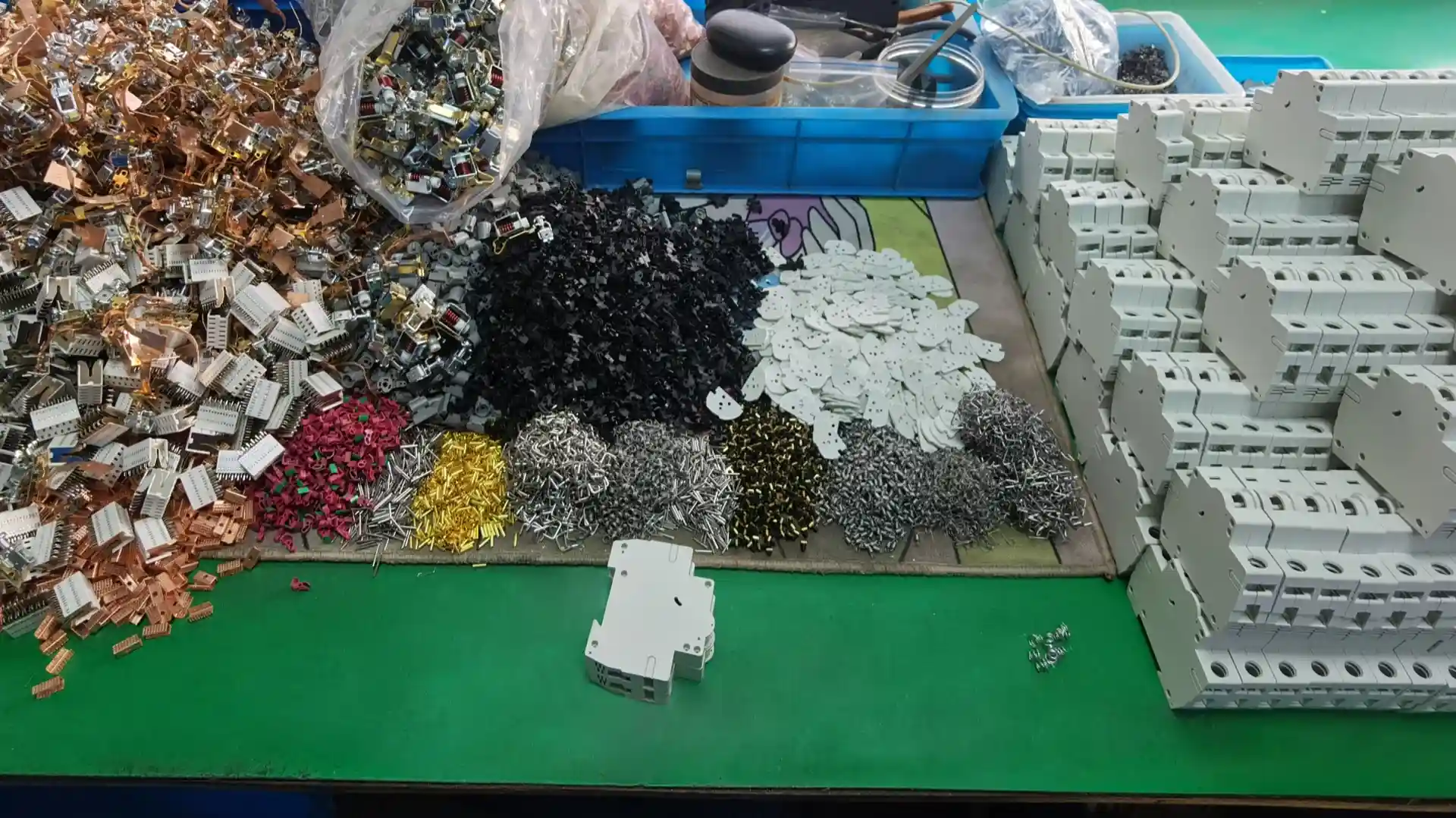 Busy green workbench in a circuit breaker manufacturing facility, covered with components for miniature circuit breakers (MCBs). In the foreground are scattered small white plastic MCB casings and bases. Behind them are organized piles of tiny parts: shiny copper contacts and coils, black plastic insulators or arc chutes, red and green plastic pieces, silver screws and rivets, gold-colored metal components, gray stamped metal plates, and heaps of assembled copper contact mechanisms. Blue plastic bins, tools, wires, and bags of additional parts are visible in the background on the green work surface.