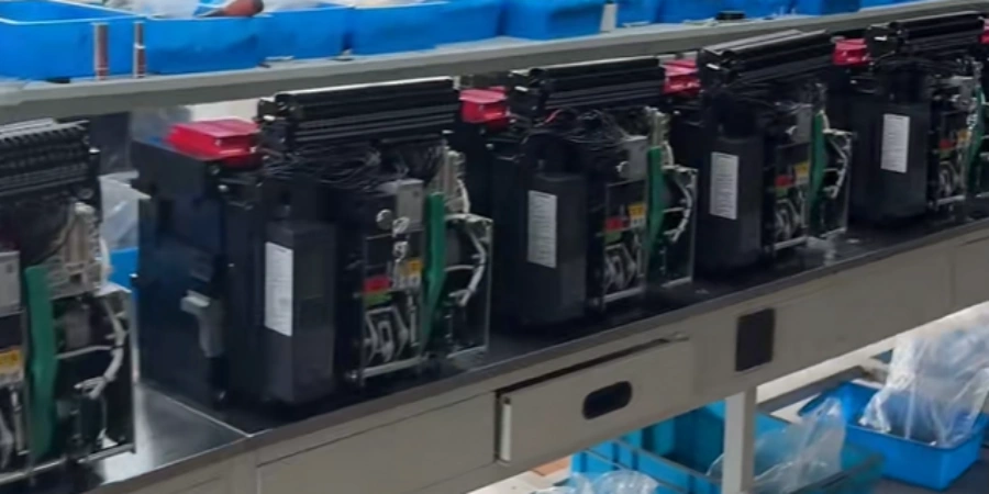 Conveyor belt production line in a factory showing a row of partially assembled large air circuit breakers (ACBs) moving along a metal conveyor, each unit in black housing with red top covers or handles, black rectangular bodies, visible internal green and yellow wiring, copper components, mechanical linkages, and labels or stickers on the sides, units mounted upright with various connectors and parts exposed, blue plastic bins overhead holding components, additional machinery and wiring visible on the left, typical industrial setup for low-voltage electrical switchgear assembly under bright factory lighting.