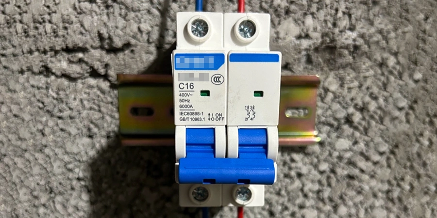 Close-up photograph of a double-pole miniature circuit breaker (MCB) clipped onto a metal DIN rail bracket against a rough gray concrete wall, the device has a white plastic body with blue toggle levers in the ON position, blue labeling strip across the top marked 'C16', green indicator windows showing status, voltage rating 400V~ 50Hz, breaking capacity 6000A, compliance markings for IEC60898-1 and GB/T 10963.1 standards, red, blue, and black wires connected to the top terminals, screw terminals at the bottom also wired, typical residential or commercial electrical protection device mounted vertically.