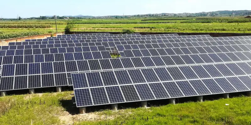 Large-scale ground-mounted solar panel installation on open land under a clear blue sky. Hundreds of dark blue photovoltaic panels are arranged in neat rows and tilted at an angle on metal support frames with concrete footings. The array is surrounded by green grassy fields, corn or crop fields in the background, distant hills, scattered trees, and a small village with houses visible on the horizon, highlighting a renewable energy project in a rural agricultural area.
