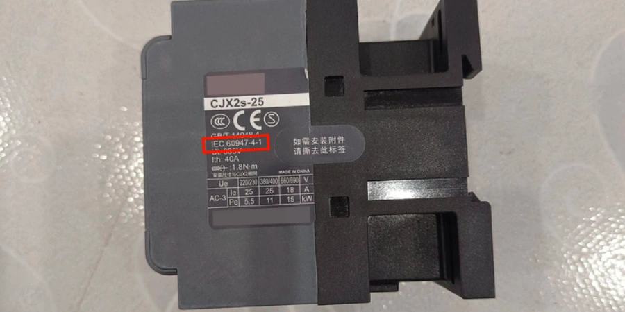 Close-up view of a gray plastic AC contactor (model CJX2s-25) with its side label visible, featuring black text on a white background including the highlighted standard 'IEC 60947-4-1', CCC and CE certification marks, technical ratings such as Ith 40A, Ue 220/380/660V, AC-3 utilization category with Ie 25A/18A and Pe 5.5/11/15 kW, and a note in Chinese about removing the label if auxiliary attachments are needed, mounted next to black mounting brackets or frame on a light surface, typical of industrial electromagnetic contactors used in motor control circuits.