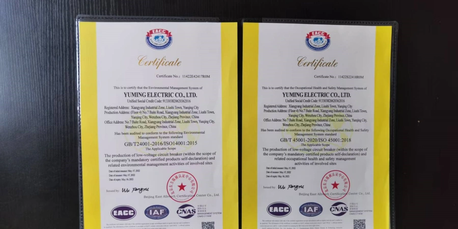Two official certificates in English are displayed side-by-side in a black folder against a dark background. The left page is an ISO 14001:2015 Environmental Management System certificate, and the right page is an ISO 45001:2018 Occupational Health and Safety Management System certificate. Both documents are issued to Yuming Electric Co., Ltd., featuring yellow side banners, red and blue EACC logos, and official accreditation stamps from IAF and CNAS at the bottom.