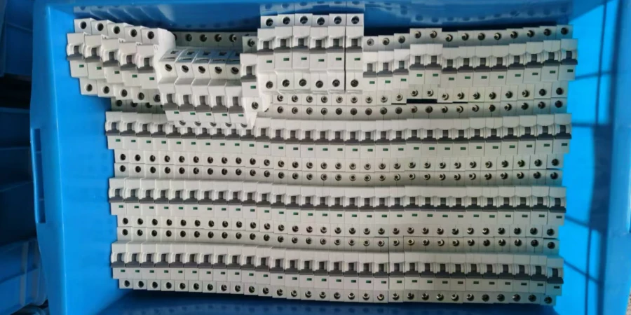 Large blue plastic storage bin filled with hundreds of white miniature circuit breakers (MCBs) stacked tightly in neat rows and layers. Each MCB has multiple screw terminals with circular openings for wiring, green marking strips or indicators on top, compact rectangular plastic bodies, and some units arranged in stepped piles at the top for easy counting or picking in a manufacturing or warehouse environment.