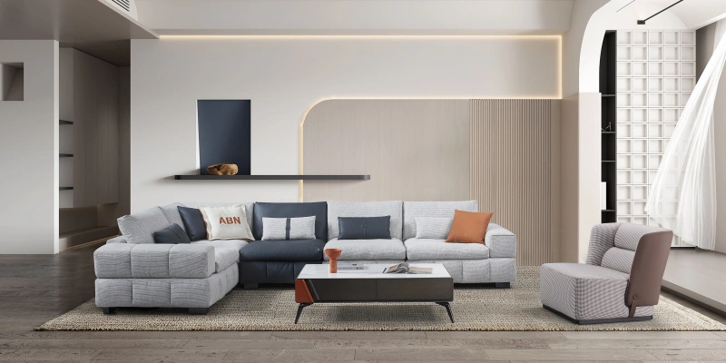 Spacious contemporary living room featuring a large light gray L-shaped sectional sofa with textured fabric upholstery, accented by navy blue and orange throw pillows including one with ‘ABN' text. The sofa sits on a neutral beige area rug over dark wood flooring. In front is a low rectangular dark gray coffee table with a slim vase and magazines. Behind the sofa is a textured beige wall with vertical wood slats and curved LED backlit niches creating warm ambient lighting. A floating black shelf holds a decorative object, a tall navy blue abstract artwork hangs above, and a matching gray armchair with ottoman is positioned to the side. The room has clean lines, neutral tones, and subtle modern elegance.
