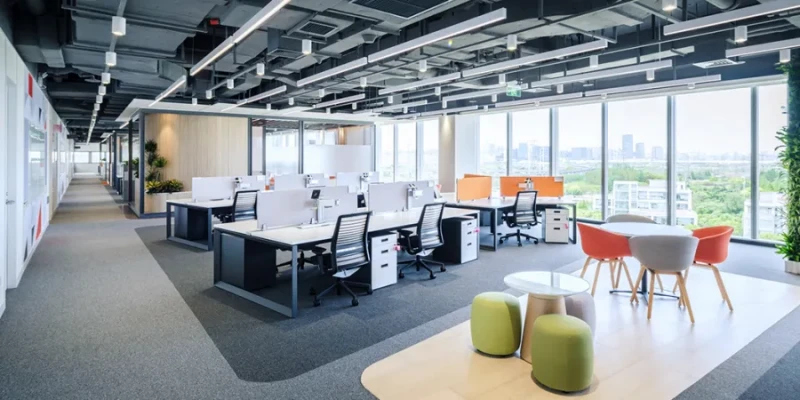 Spacious contemporary open-plan office on a high floor, featuring long white desks arranged in rows with ergonomic black and white office chairs. Workstations are equipped with large monitors and divided by low partitions. The space includes colorful casual seating areas with orange, gray, and red round tables, paired with matching chairs and green cylindrical stools. Exposed dark gray ceiling with linear LED lights and air ducts, glass-walled meeting rooms along one side, potted plants and wooden accents, and floor-to-ceiling windows offering a panoramic view of a city skyline, green landscape, and distant buildings under a bright sky.