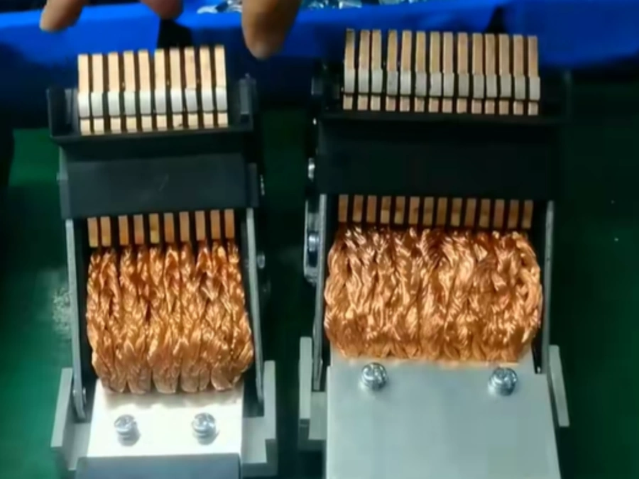 Close-up view of two moving contact units for air circuit breakers, each showing dense bundles of shiny copper braided strands or laminated copper strips coiled and compressed into thick, wavy orange-brown contact blocks, topped with neatly aligned rows of copper bars or fingers for electrical connection, held securely in jigs with visible bolts at the base, hands partially visible above adjusting or inspecting the components, set on a blue-covered table in an industrial manufacturing setting with green floor visible, typical of high-current electrical switchgear production.