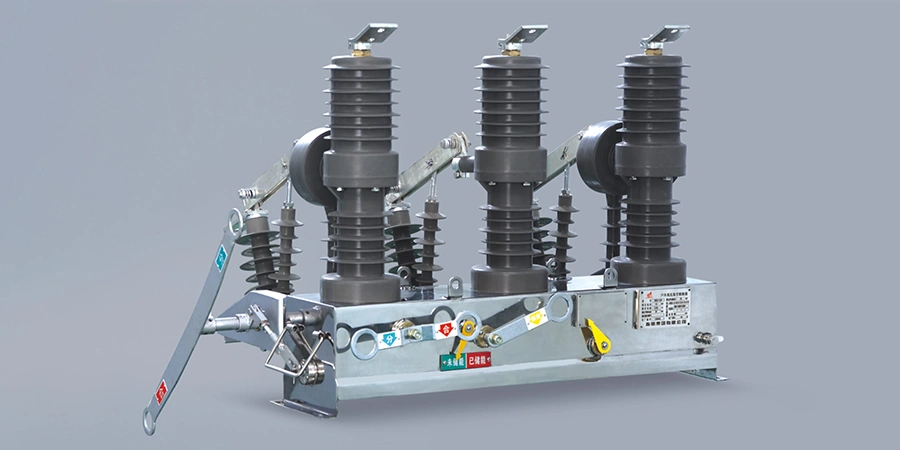 A three-pole outdoor vacuum circuit breaker designed for high-voltage applications, featuring three tall gray ceramic or polymer insulators with deep ribbed skirts for each phase, topped with metal connectors and arcing horns. The unit has a sturdy gray metal base frame with mounting feet, visible operating linkage arms, a large manual operating lever on the left side, and various control rods and mechanisms. A silver control box or label plate at the bottom includes warning stickers, grounding points, and technical information plates. The breaker is shown against a neutral gray background, highlighting its robust construction for substation or distribution use.