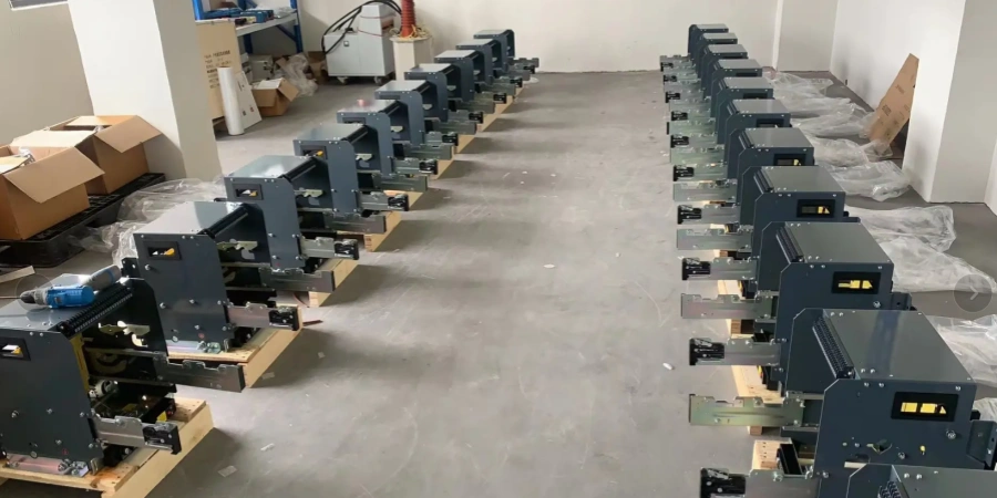 Long row of identical gray draw-out type air circuit breaker (ACB) chassis or cassettes lined up in a straight line across a spacious concrete factory floor. Each unit is mounted on a wooden pallet, featuring a metal frame with sliding rails, visible top terminal blocks with multiple connection points, black operating mechanisms, yellow and black warning labels or indicators on the front, and various internal components like linkages and mounting brackets exposed. The breakers are partially assembled or ready for installation, surrounded by cardboard boxes, plastic wrapping, tools, and industrial equipment in the background of a clean workshop environment.