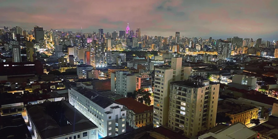 Panoramic cityscape of São Paulo at dusk or early night, dense urban skyline filled with countless illuminated high-rise buildings and apartment blocks, prominent pinkish-purple lit tower standing out in the center, vibrant mix of warm yellow, white, red, and blue lights from windows and streets, foreground showing nearby rooftops and mid-rise structures with some greenery, dramatic cloudy sky in shades of orange, pink, and gray transitioning to night, bustling metropolitan atmosphere captured from an elevated viewpoint.