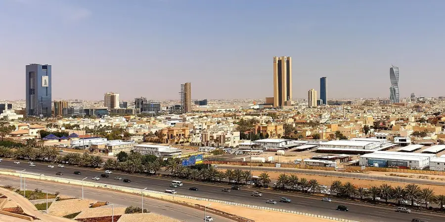 Wide aerial view of modern Riyadh under a clear blue sky, featuring a dense urban landscape with clusters of mid-rise and high-rise buildings in beige, tan, and white tones, prominent modern skyscrapers including a tall twisted tower on the right, twin golden high-rises in the center, a sleek blue glass tower, and a large blue rectangular building with signage on the left, wide multi-lane highway in the foreground lined with palm trees carrying heavy traffic of cars, low-rise residential and commercial areas filling the midground, sandy desert-like surroundings visible in the distance.
