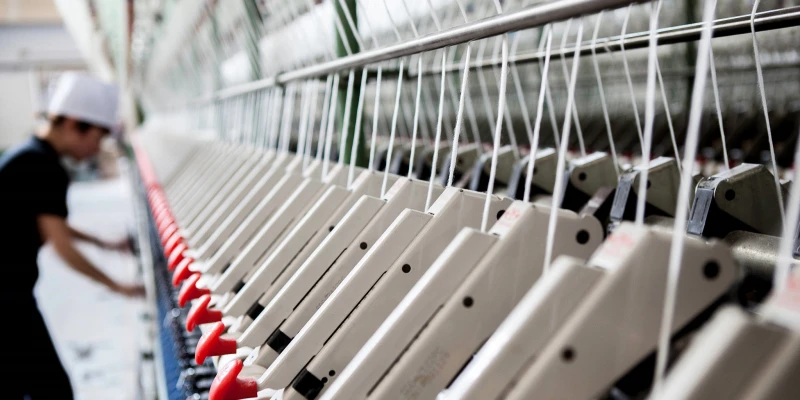 Close-up view of a modern industrial spinning machine in a textile factory. Long rows of white yarn bobbins or spindles are aligned along metal frames, with thin white threads running vertically under tension. Red and white shuttle or flyer mechanisms guide the yarn, while a worker in a black shirt and white cap adjusts the machine in the background. The setup features shiny chrome bars, green accents, and bright factory lighting highlighting the precise, high-speed yarn production process.