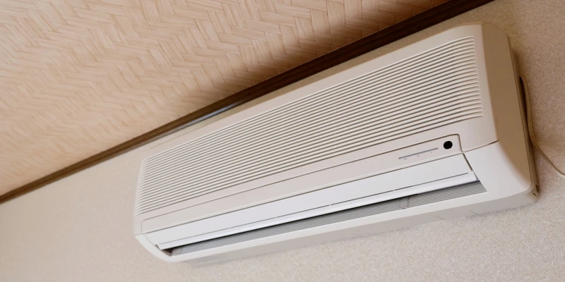 A sleek beige wall-mounted split-system air conditioner indoor unit installed high on a light-colored ceiling with a textured bamboo or herringbone pattern. The unit has a clean horizontal design with fine horizontal louvers for air distribution, a small black sensor or indicator light in the center, and a subtle control panel on the right side. It blends neatly into the room's warm neutral tones, mounted below a dark wooden trim strip.