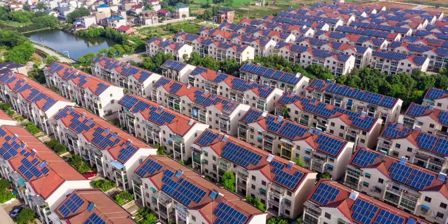 Drone aerial photograph of a densely packed residential village in rural China, featuring rows of multi-story houses with bright red-tiled roofs completely covered in blue solar photovoltaic panels. The neighborhood is surrounded by green trees, farmland, and a small pond or river, showcasing large-scale adoption of rooftop solar energy in a suburban-rural setting under clear daylight.