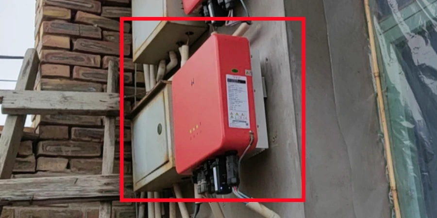 Red rectangular solar inverter mounted on a brick and concrete wall, highlighted in a red rectangle. The unit has a green LED indicator light, white label with text and warnings, black cables and connectors at the bottom, surrounded by wooden supports, white pipes, and green construction netting.