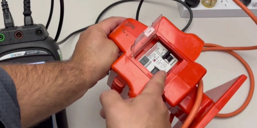 Technician holding a bright red portable power strip with its clear plastic lid open, showing the internal residual current device (RCD) and CE marking, connected to orange extension cables.