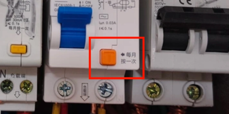 Close-up of a white residual current device (RCD) showing an orange test button labeled 'Press once per month' highlighted in a red box, with blue switch lever and wiring terminals visible.