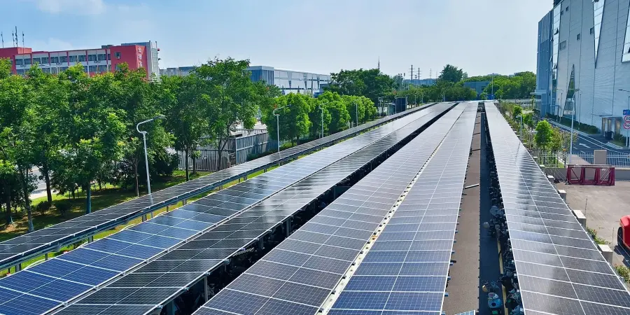 Long rows of dark blue solar photovoltaic panels mounted on metal frames, extending in parallel lines across an open area next to a large modern factory building with red and gray sections. Surrounded by green trees, clear blue sky with some clouds, street lamps, and distant urban structures.
