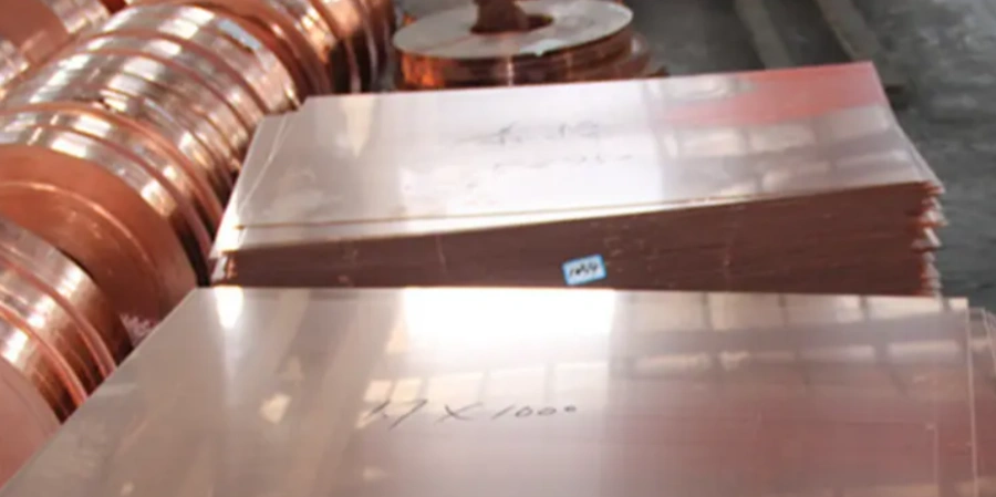 Stack of polished T2 copper sheets/plates in the foreground with handwritten markings like '17 X 1000' and Chinese characters, surrounded by multiple reels of copper coil in an industrial storage area.