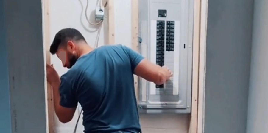 Side view of an electrician in a teal blue t-shirt standing to the side of an open electrical distribution panel mounted in a wooden frame on a construction wall. His body is positioned sideways, face turned away from the panel and camera for safety, arms extended forward as he reaches in to flip or close one of the circuit breakers in the vertical row inside the white metal enclosure, colorful wires visible along the sides, typical safety practice when operating live or high-current breakers in electrical installation work.